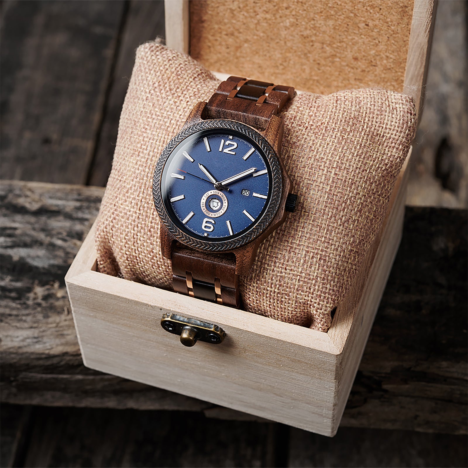 A sophisticated men's wooden watch featuring a blue dial, date display, textured bezel, and rose gold-accented band, presented in a wooden box with a burlap pillow against a rustic wood background to emphasize its elegant and timeless packaging.