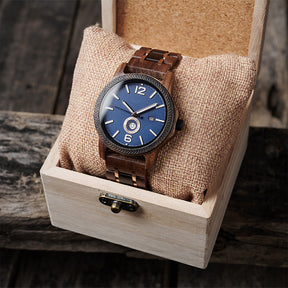 A sophisticated men's wooden watch featuring a blue dial, date display, textured bezel, and rose gold-accented band, presented in a wooden box with a burlap pillow against a rustic wood background to emphasize its elegant and timeless packaging.