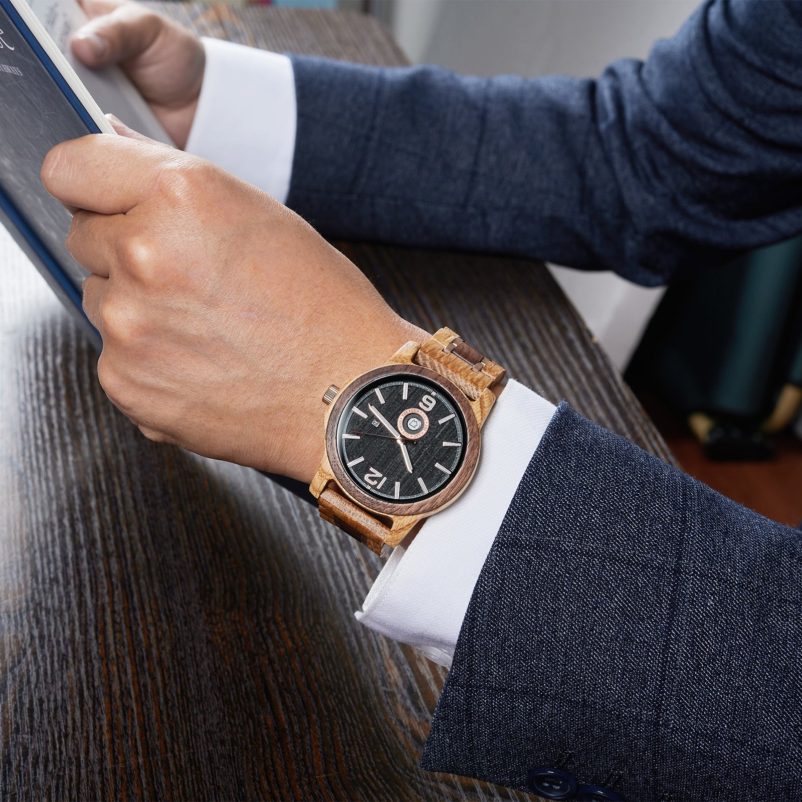 A sophisticated men's wooden watch featuring a dark gray dial, showcased on a wrist wearing a navy suit while reading a book, highlighting its elegant and professional aesthetic.