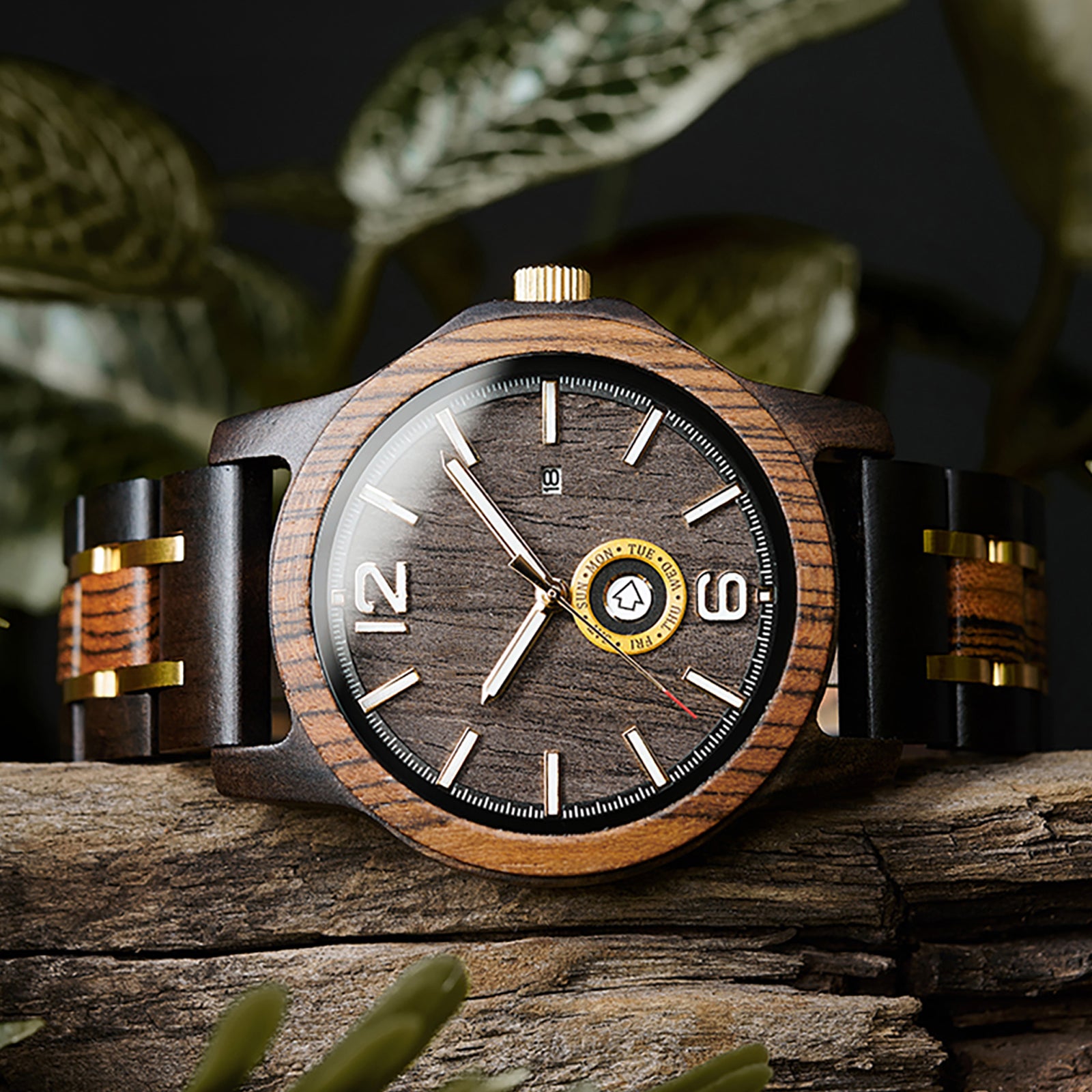 A stylish men's wooden watch featuring a dark gray dial, date and day display, natural wood grain band with gold accents, presented against a backdrop of rustic wood and green foliage to highlight its rugged yet sophisticated aesthetic.