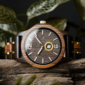 A stylish men's wooden watch featuring a dark gray dial, date and day display, natural wood grain band with gold accents, presented against a backdrop of rustic wood and green foliage to highlight its rugged yet sophisticated aesthetic.