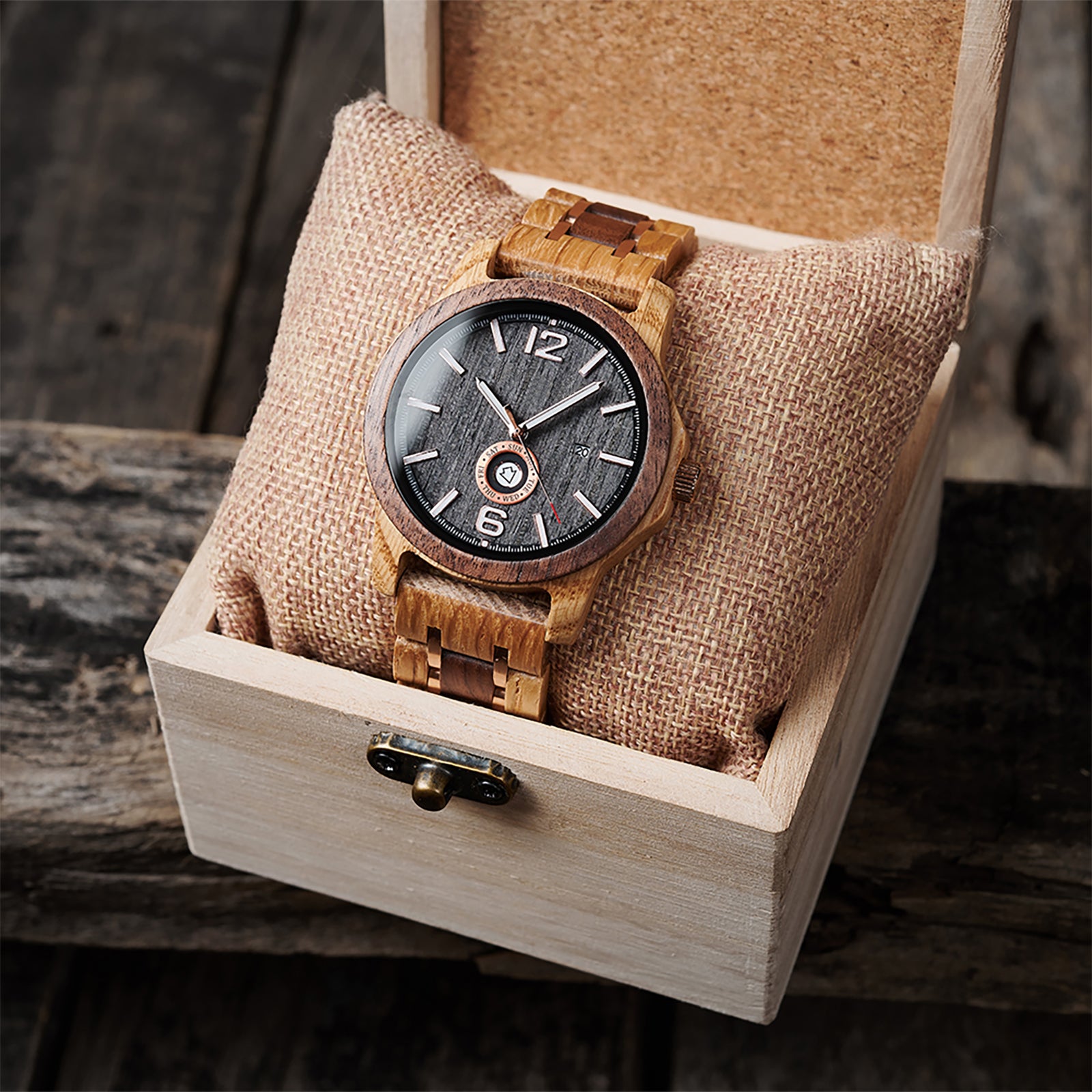 A sophisticated men's wooden watch featuring a dark gray dial, date and day display, natural wood grain band with rose gold accents, presented in a wooden box with a burlap pillow against a rustic wood background to emphasize its elegant and timeless packaging.