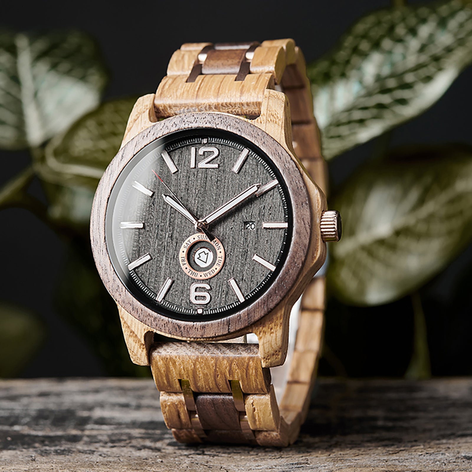 A stylish men's wooden watch featuring a dark gray dial, date and day display, natural wood grain band, presented against a backdrop of green foliage to highlight its rugged yet sophisticated aesthetic.