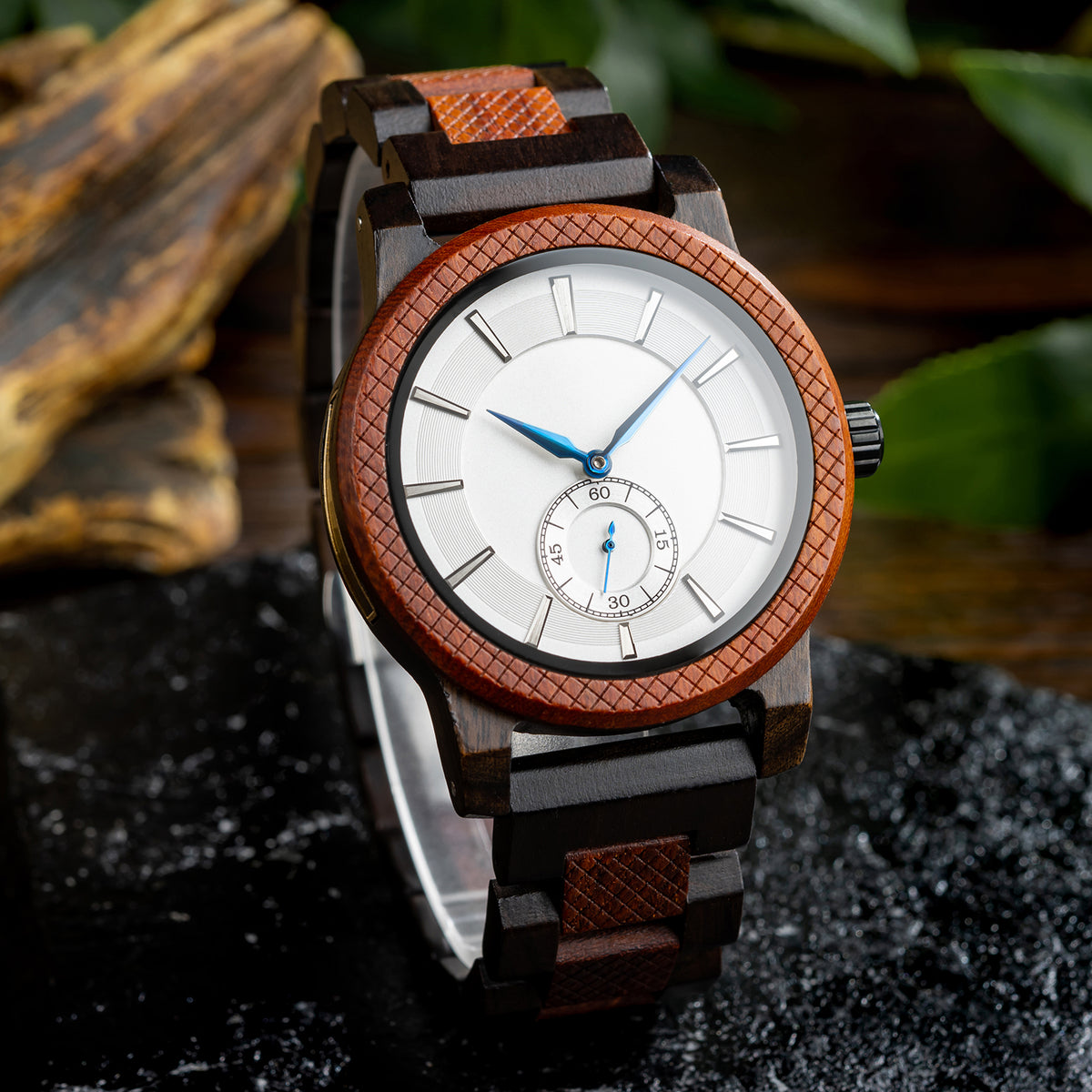 A stylish men's wooden watch featuring a white dial, sub-dial, textured brown bezel, and blue-accented hands, presented against a backdrop of rustic wood and green foliage to highlight its sophisticated and natural aesthetic.