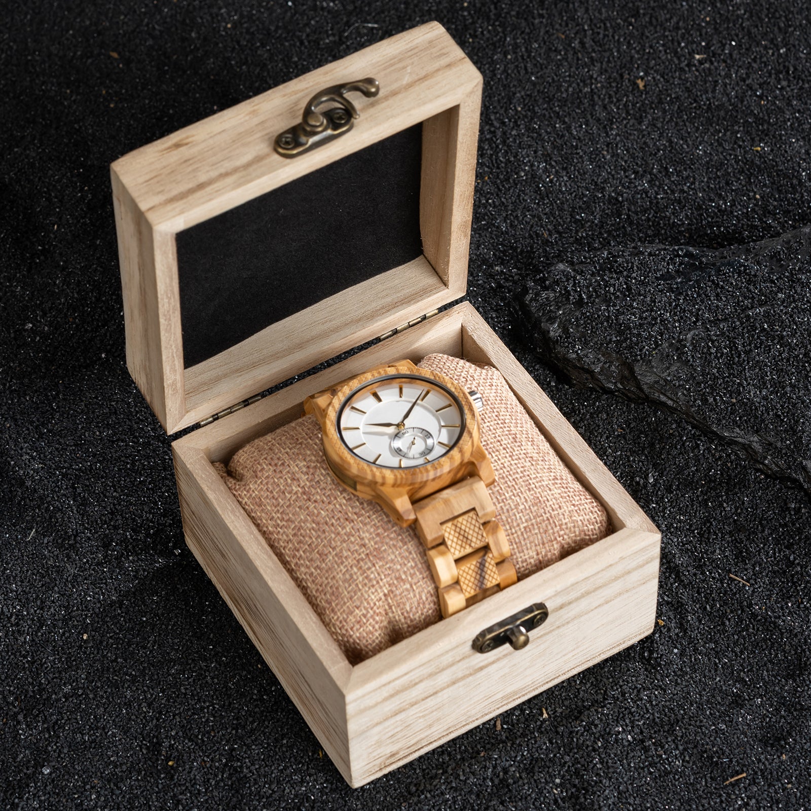 A sophisticated men's wooden watch featuring a white dial, sub-dial, textured light brown bezel, and gold-accented details, presented in a wooden box with a burlap pillow against a black pebbles background to emphasize its elegant and natural packaging.