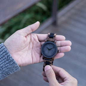 A rugged men's watch featuring a gray stone dial, wooden and black accent band, and industrial design elements, showcased in a hand against an outdoor backdrop with greenery and wooden structures to highlight its masculine and natural aesthetic.