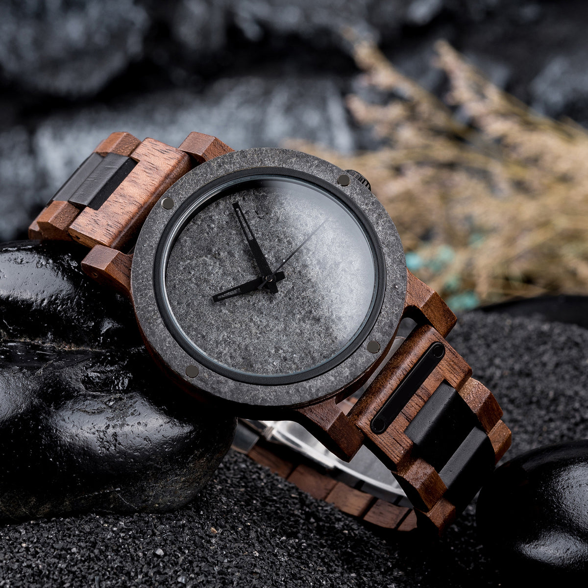 A rugged men's watch featuring a gray stone dial, wooden and black accent band, and industrial design elements, presented on black rocks amidst a dark, textured background to highlight its masculine and natural aesthetic.