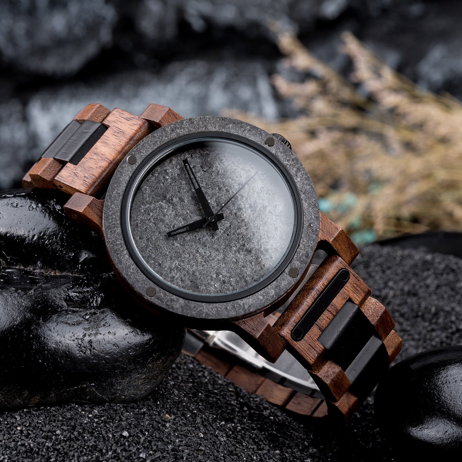 A rugged men's watch featuring a gray stone dial, wooden and black accent band, and industrial design elements, presented on black rocks amidst a dark, textured background to highlight its masculine and natural aesthetic.
