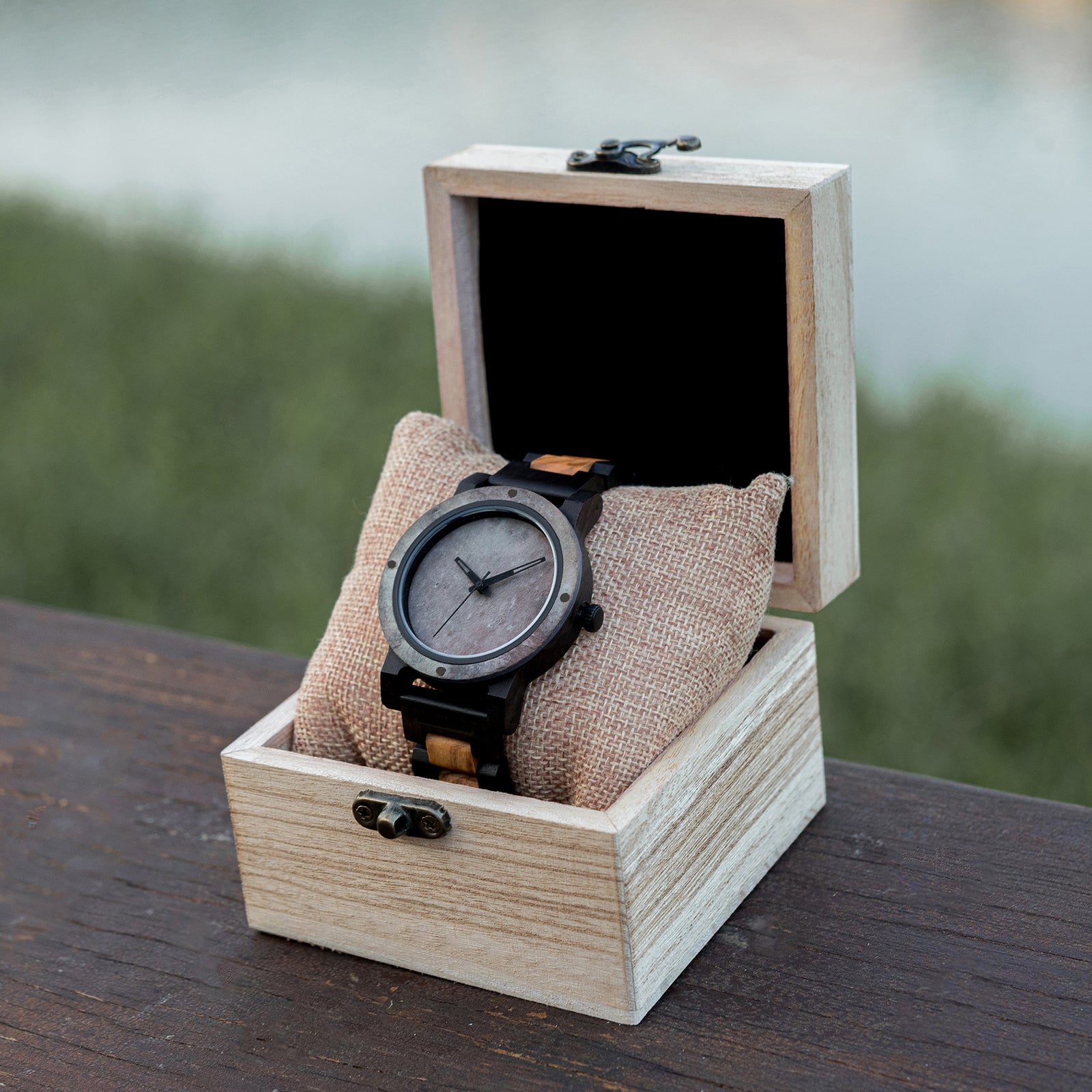 A rugged men's watch featuring a gray stone dial, two-tone wooden band with dark and light wood accents, presented in a wooden box with a burlap pillow amidst an outdoor backdrop of greenery and a wooden surface to highlight its masculine and natural aesthetic.