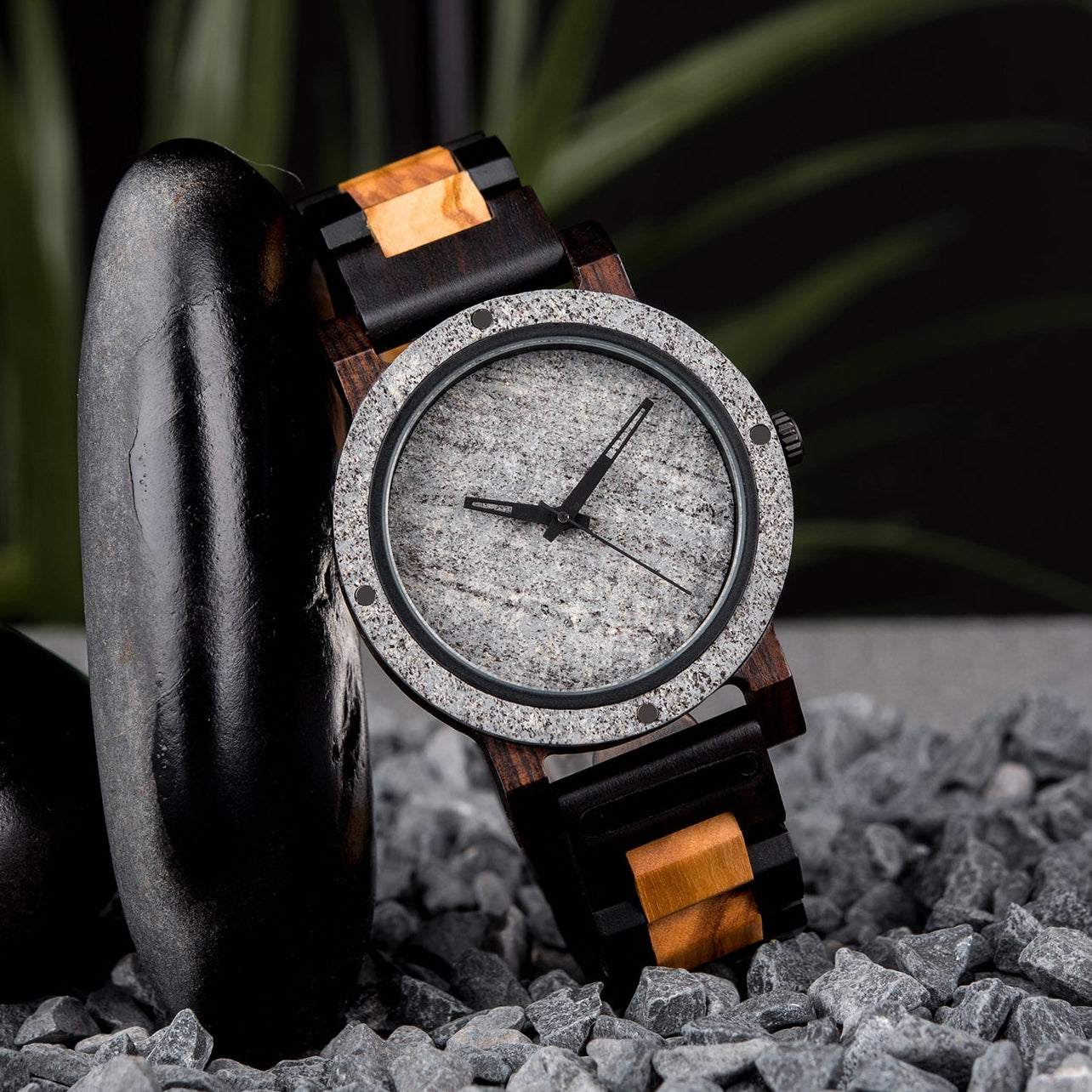 A rugged men's watch featuring a gray stone dial, two-tone wooden band with dark and light wood accents, presented on gray rocks amidst a dark, textured background with green foliage to highlight its masculine and natural aesthetic.
