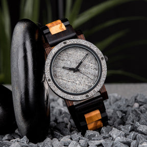 A rugged men's watch featuring a gray stone dial, two-tone wooden band with dark and light wood accents, presented on gray rocks amidst a dark, textured background with green foliage to highlight its masculine and natural aesthetic.