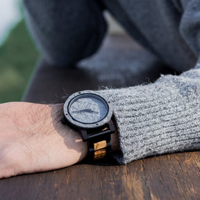 A rugged men's watch featuring a gray stone dial, two-tone wooden band with dark and light wood accents, showcased on a wrist with a gray knit sweater, resting on a wooden table to highlight its masculine and natural aesthetic.