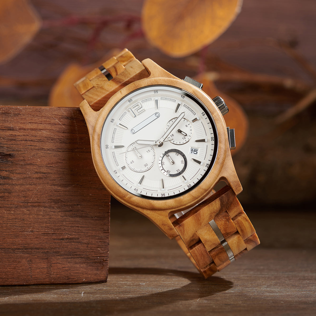 A wooden chronograph watch with a white dial, multiple sub - dials, date "15" display, and a strap made of wooden links with metal accents, positioned on a wooden surface with blurred natural elements in the background.