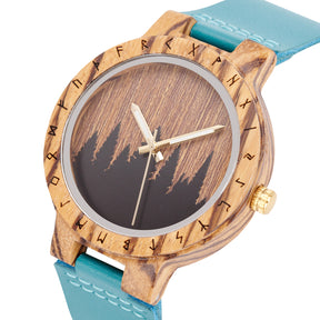 A close-up of a unique wooden watch with a runic symbol bezel, forest silhouette dial, turquoise leather band, and gold-tone hands, highlighting the natural wood texture against a white background.