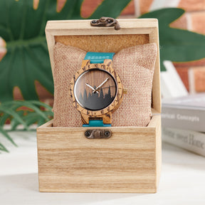 A unique wooden watch with a runic symbol bezel, forest silhouette dial, and turquoise leather band, nestled on a burlap pillow inside a wooden box, with a blurred background of green foliage and books.