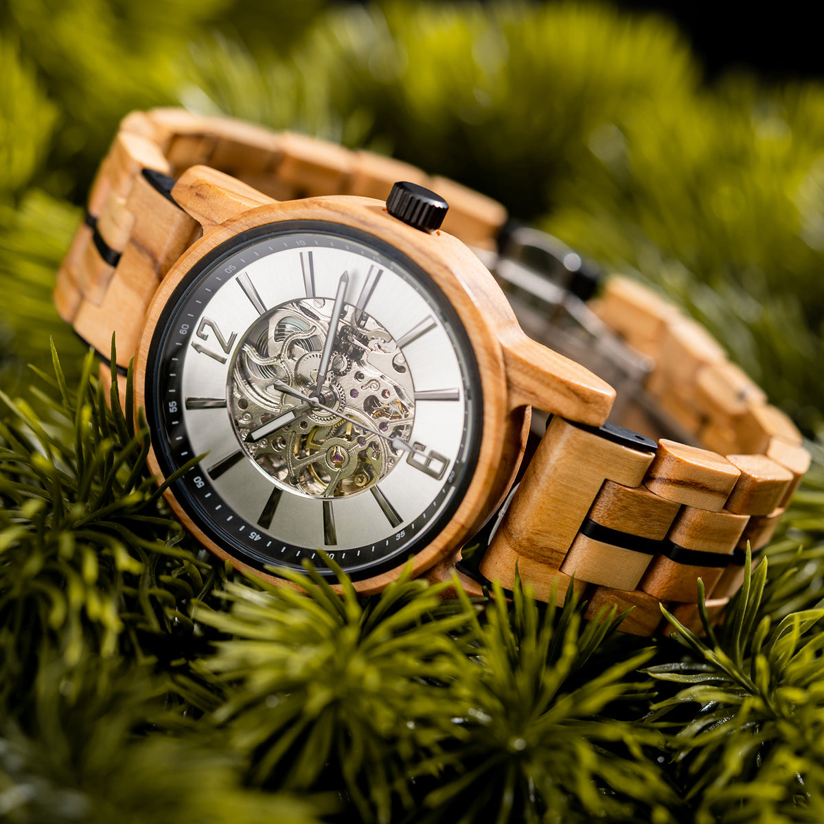 A wooden wristwatch with a silver dial, full skeleton movement, and black-accented wooden band, placed on green pine foliage.