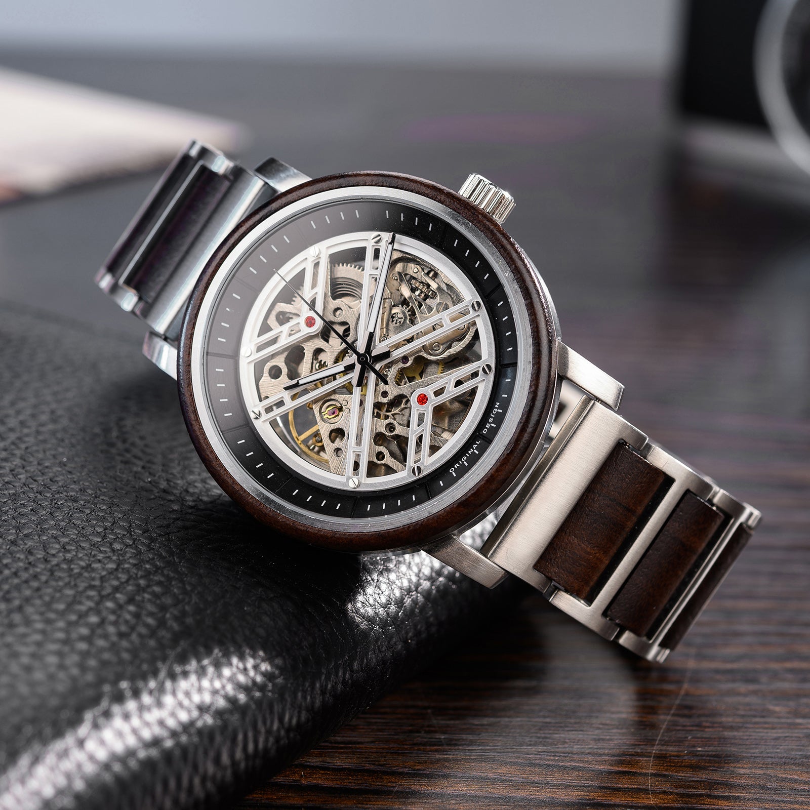 A men's skeleton mechanical watch featuring a silver metal and wooden case and band, showcasing intricate internal mechanisms with red accents through a skeleton dial, positioned on a black leather surface with a wooden background.