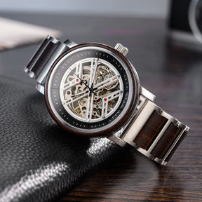 A men's skeleton mechanical watch featuring a silver metal and wooden case and band, showcasing intricate internal mechanisms with red accents through a skeleton dial, positioned on a black leather surface with a wooden background.