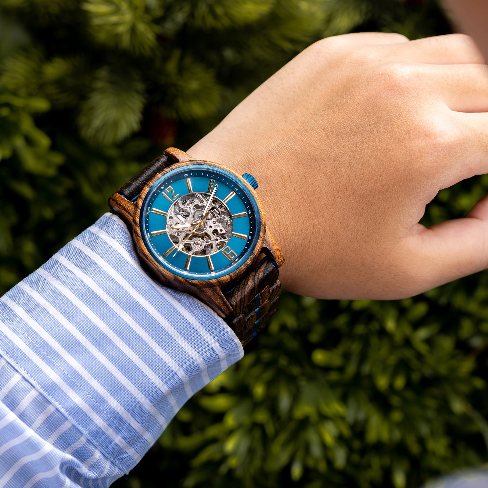 A person wearing a teal dial full skeleton wooden wristwatch with blue accents, paired with a light blue striped shirt, against a green foliage background.