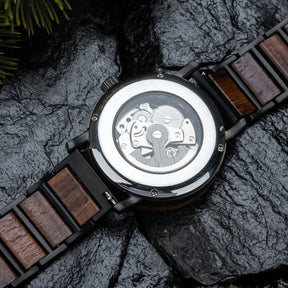 Back view of a mechanical wristwatch with visible gears, wood and black metal combined band, placed on dark rocky surface with green pine foliage accent.