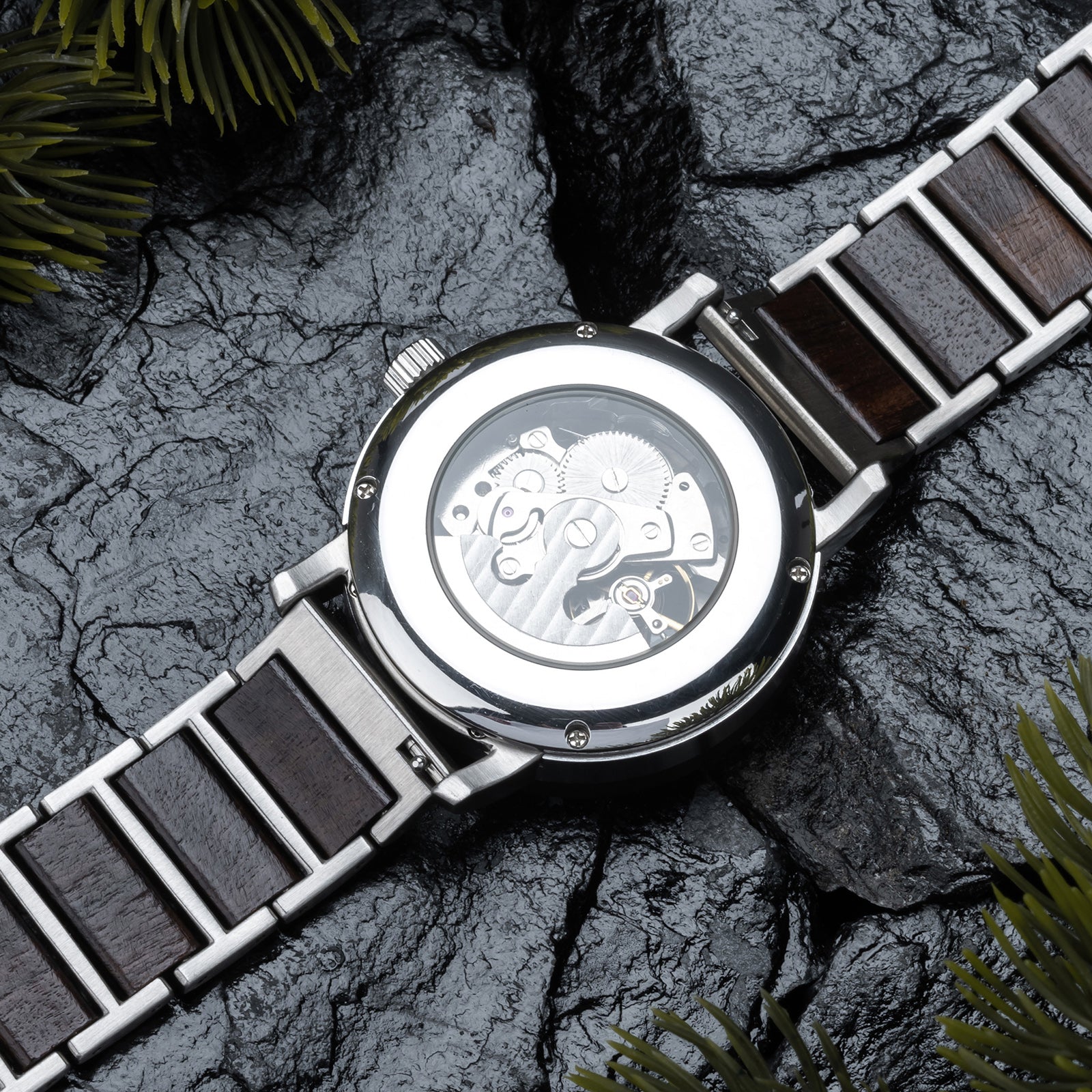 Back view of a mechanical wristwatch with visible gears, wood and metal combined band, placed on dark rocky surface with green foliage accents.