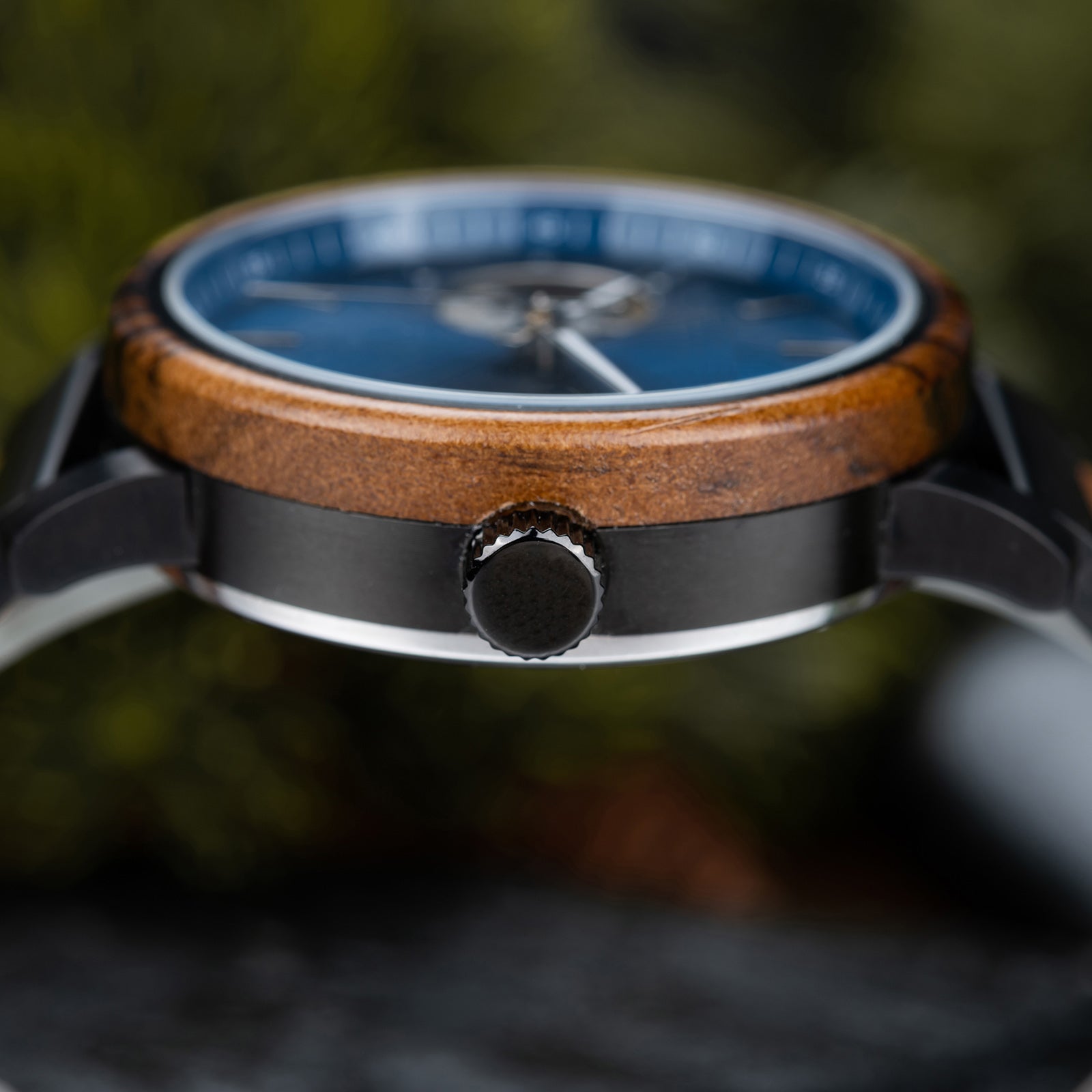 Close-up of a wristwatch's crown, wooden bezel, and blue dial, with a blurred green foliage background.