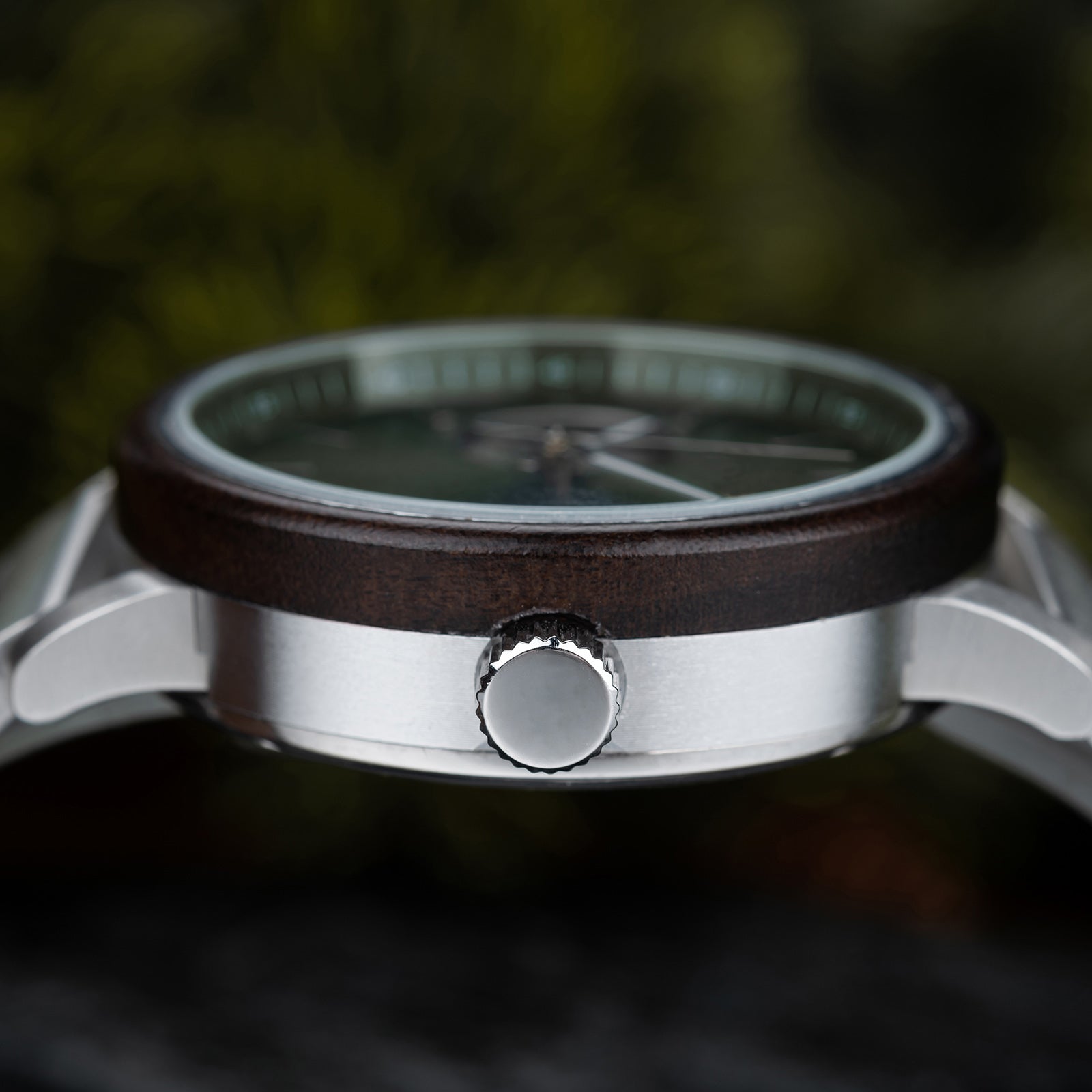 Close-up of a wristwatch's crown, wooden bezel, and green dial, with a blurred green foliage background.
