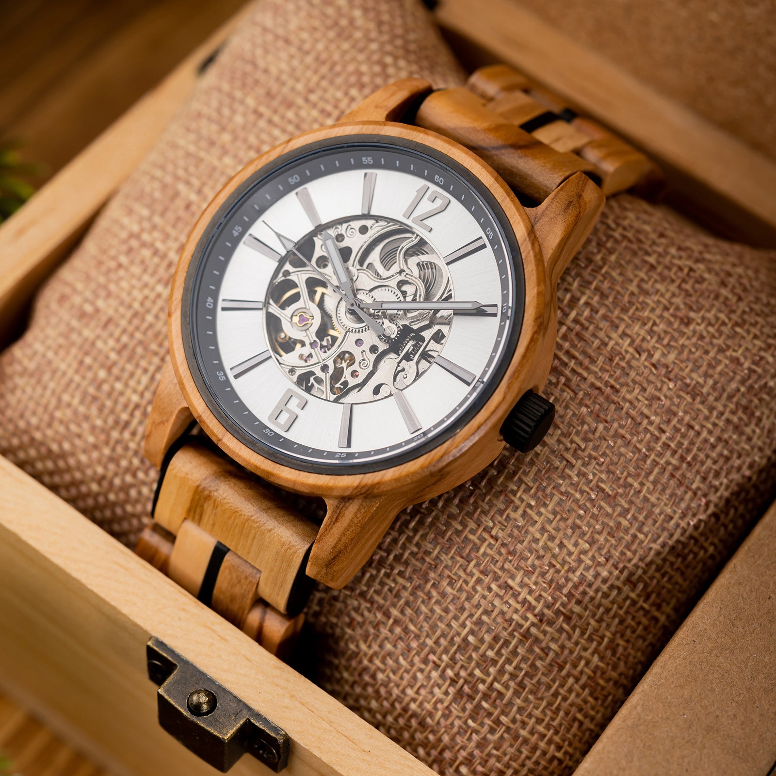 A wooden wristwatch with a white dial, full skeleton movement, and black accents, placed inside a wooden box with burlap padding.