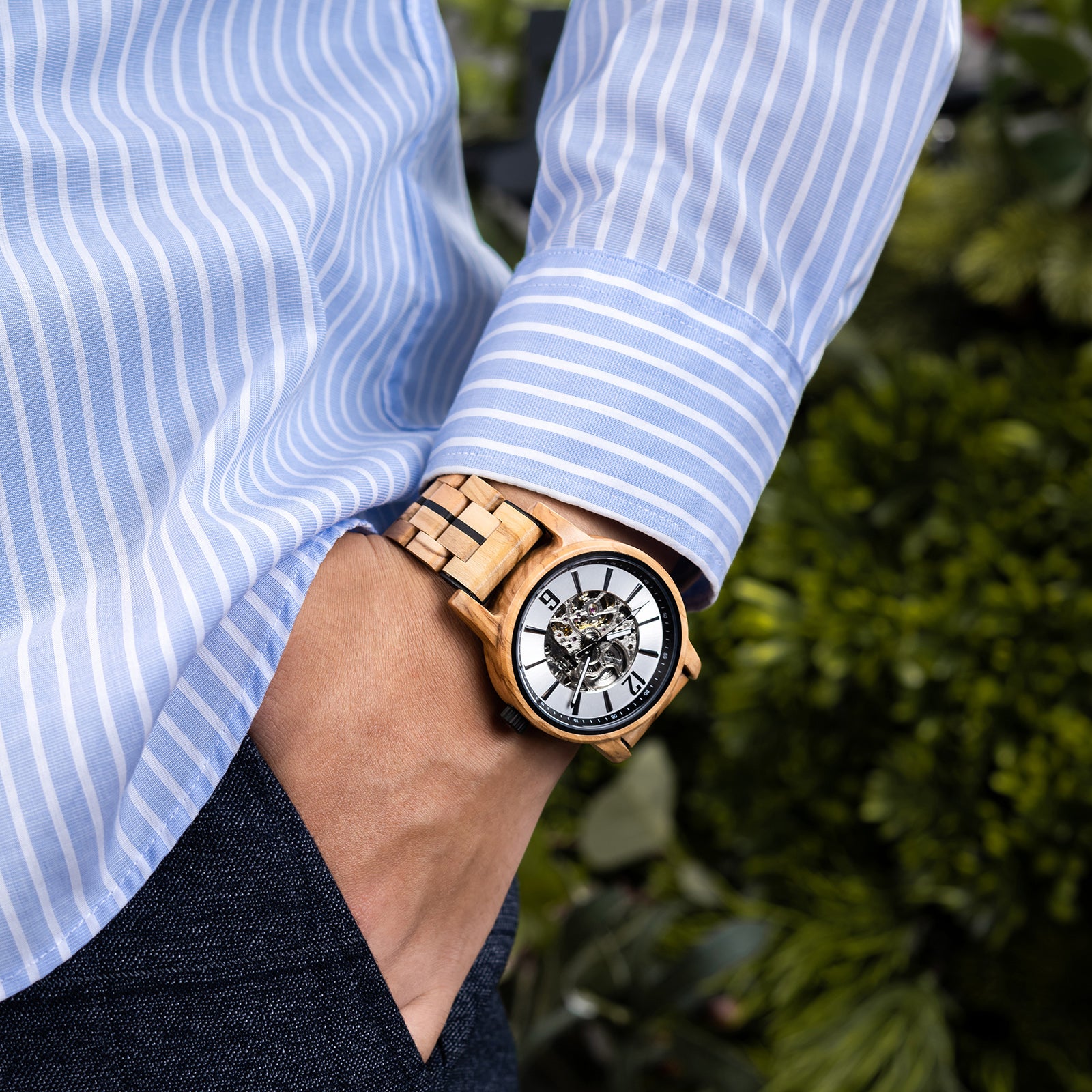 A person wearing a white dial full skeleton wooden wristwatch with black accents, paired with a light blue striped shirt, against a green foliage background.