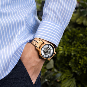 A person wearing a white dial full skeleton wooden wristwatch with black accents, paired with a light blue striped shirt, against a green foliage background.