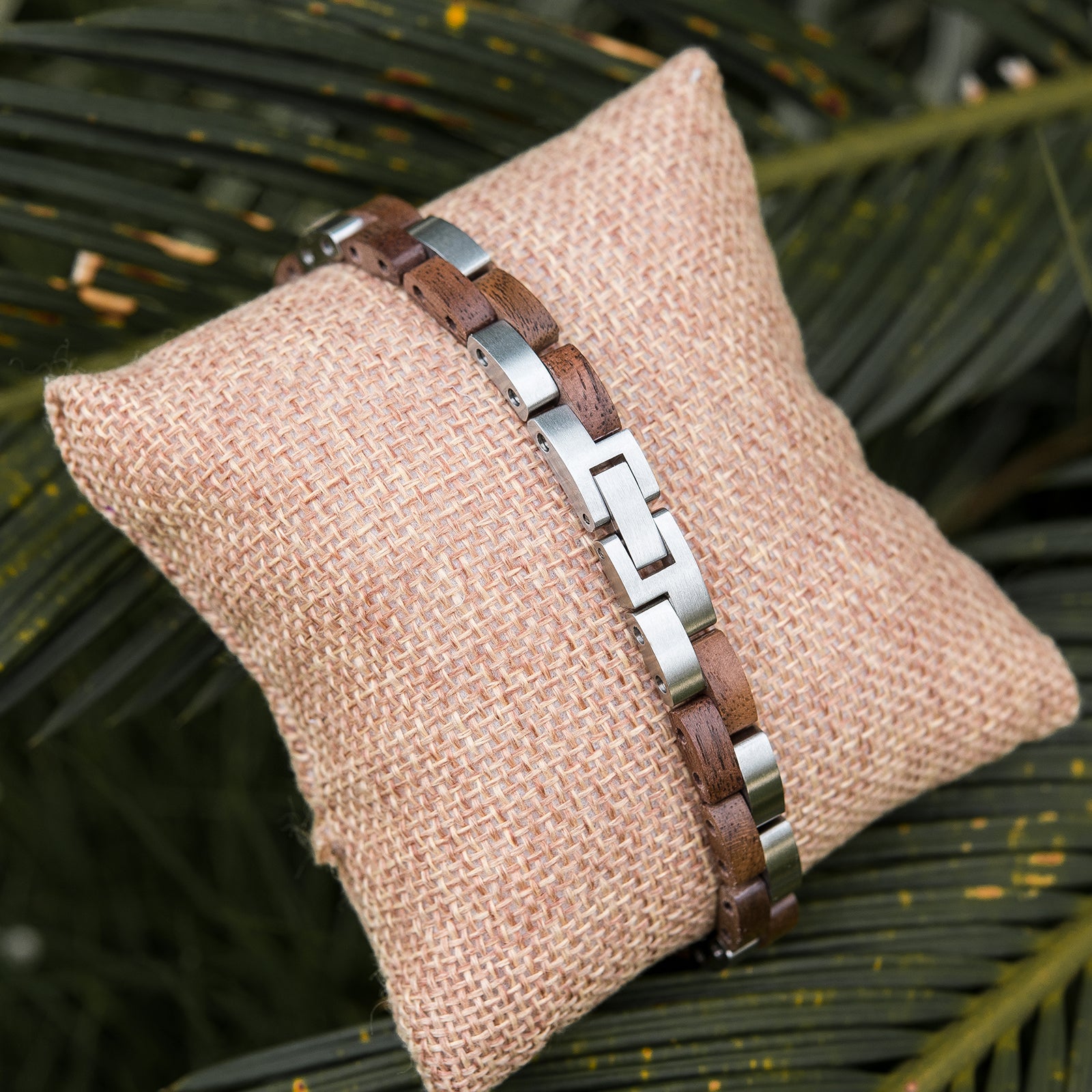 Women's wooden bracelet with stainless steel links, placed on burlap pillow amidst tropical leaves. Boho-chic, nature-inspired design, perfect for stylish, feminine accessory lovers.