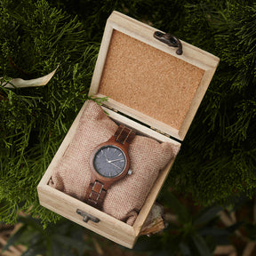 An elegant women's walnut watch presented in a wooden box with a burlap cushion, set against a backdrop of lush greenery to emphasize its natural and refined packaging.