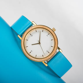 A stylish women's watch with a wooden-accented bezel, white dial, gold-tone details, and a blue leather band, placed on a blue surface against a light gray background.