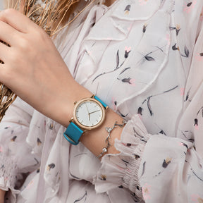 A stylish women's watch with a wooden-accented bezel, white dial, blue leather band, paired with a floral dress and a delicate bracelet, showcasing a fashionable and feminine ensemble.
