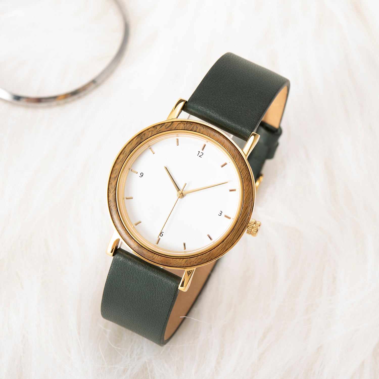 A stylish women's watch with a wooden-accented bezel, white dial, gold-tone details, and a dark green leather band, placed on a white fur surface with a subtle metallic accessory in the background.