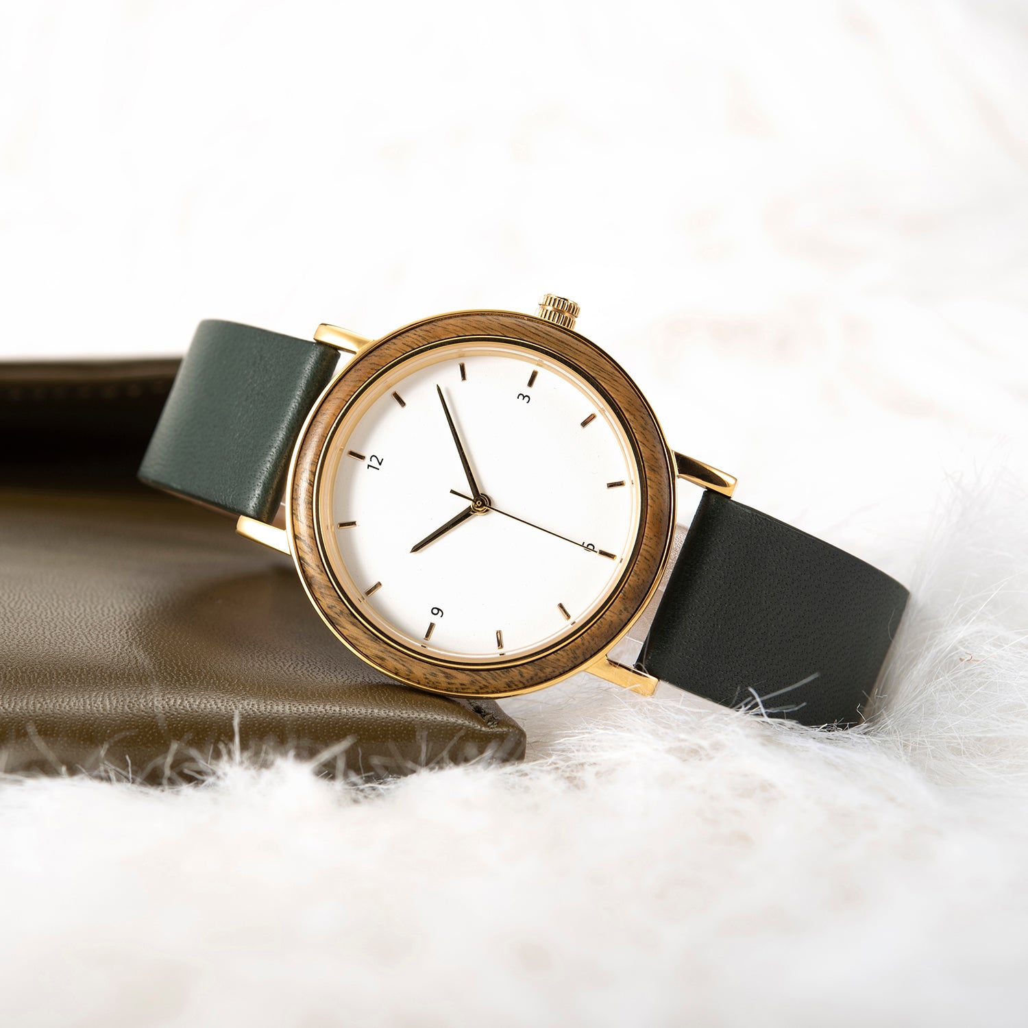 A stylish women's watch with a wooden-accented bezel, white dial, gold-tone details, and a dark green leather band, placed on a white fur surface with a brown leather accessory in the background.