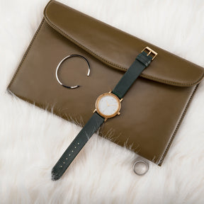 A stylish women's watch with a wooden-accented bezel, white dial, gold-tone details, and a dark green leather band, paired with an olive leather clutch and minimalist jewelry, displayed on a white fur surface.