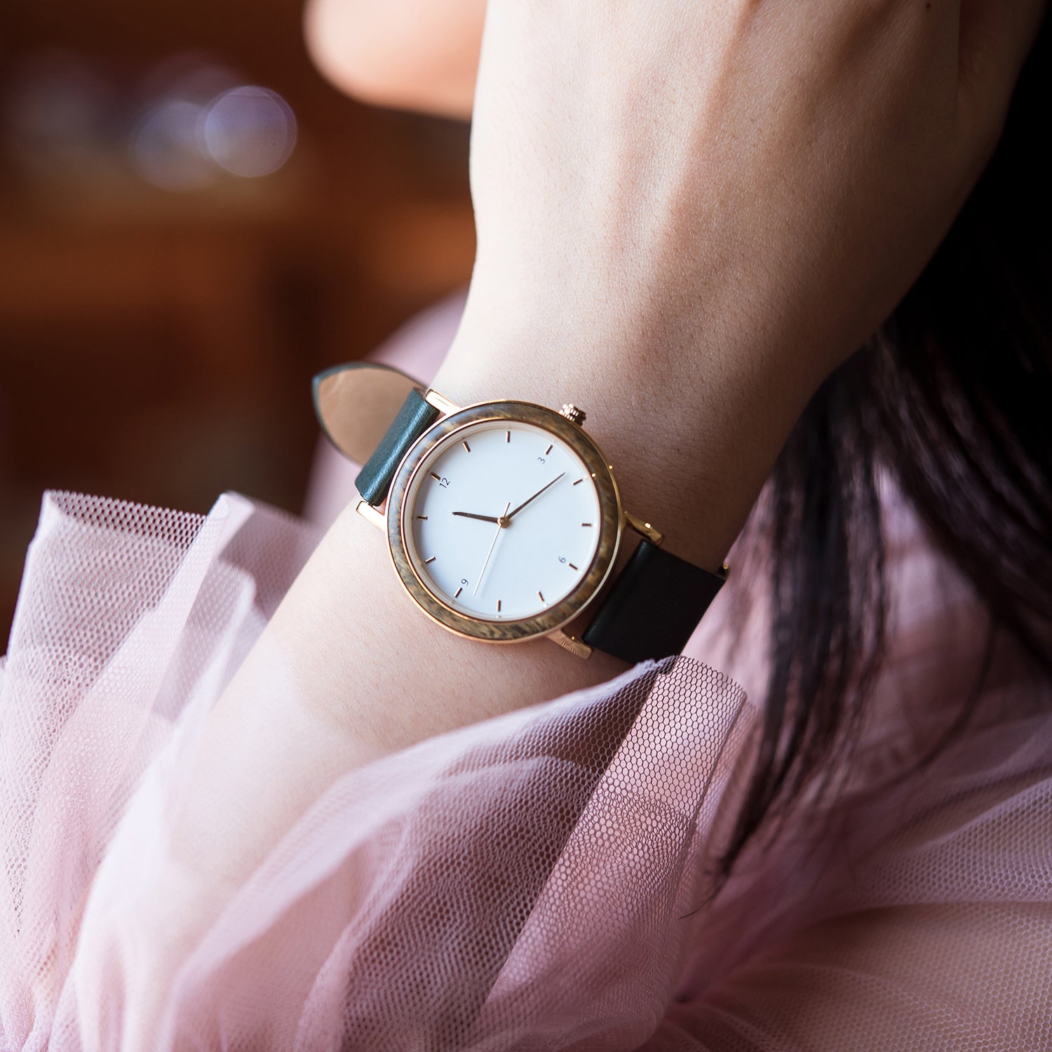 A stylish women's watch with a wooden-accented bezel, white dial, gold-tone details, and a dark green leather band, worn with a pink tulle dress, creating a feminine and fashionable look.