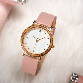 A stylish women's watch with a wooden-accented bezel, white dial, gold-tone details, and pink leather band, placed on a soft white fur surface with subtle fashion accessories in the background.