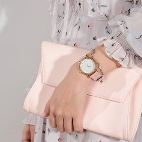 A stylish women's watch with a wooden-accented bezel, white dial, pink leather band, paired with a pink clutch and a floral dress, showcasing a fashionable ensemble.
