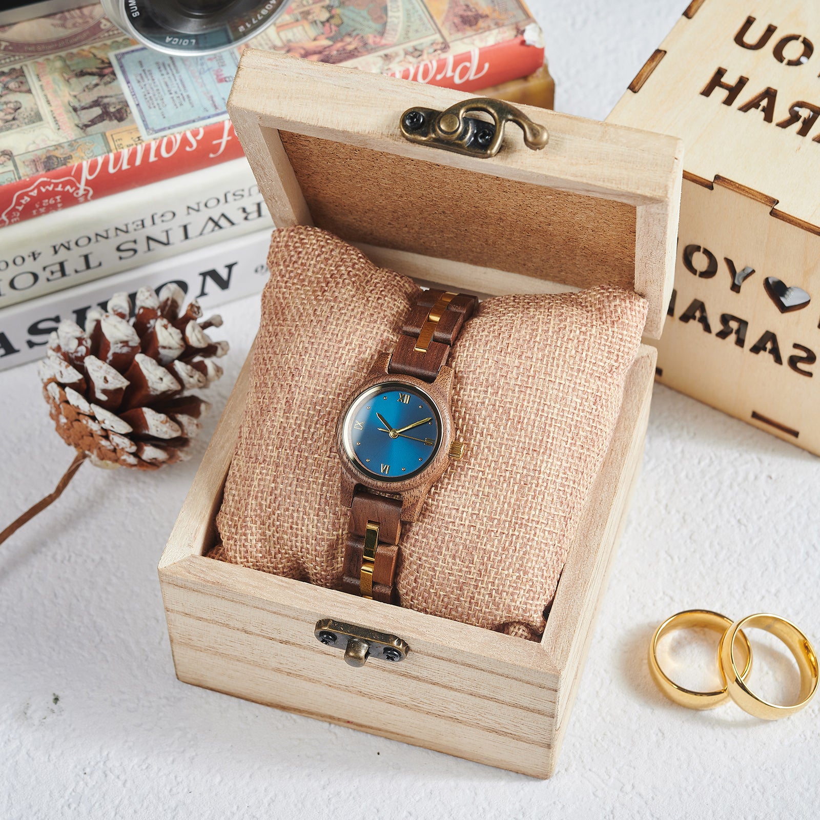 A sophisticated women's wooden watch with a blue dial and gold accents, presented in a wooden box with a burlap cushion, set against a vintage background of books, a pinecone, and gold rings to emphasize its elegant and timeless packaging.