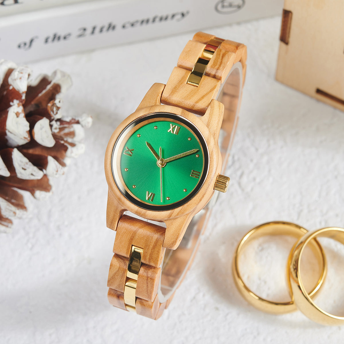 A stylish women's wooden watch with a green dial, gold accents, and Roman numerals, presented with vintage elements like a pinecone and gold rings to highlight its elegant and timeless aesthetic.