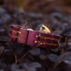 A detailed view of a women's wooden watch band featuring purple wood and gold-tone metal accents, nestled amidst dark foliage to highlight its luxurious and natural craftsmanship.

