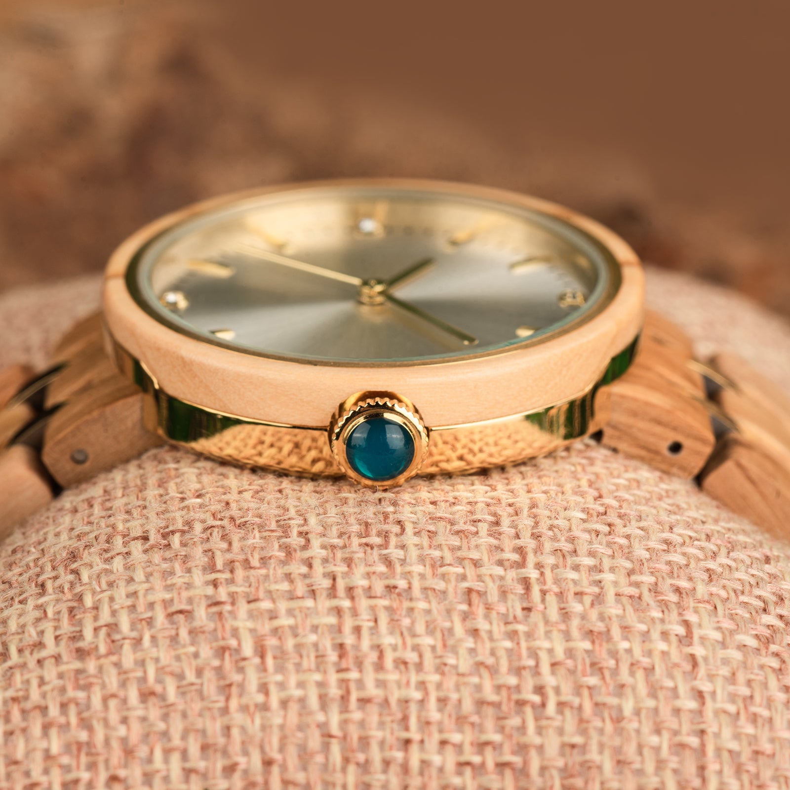 A close-up view of a women's wooden watch featuring a gold-tone dial, diamond-like details, a blue gem crown, and a two-tone wood and metal band, placed on burlap to highlight its luxurious craftsmanship and design details.