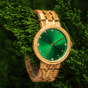 An elegant women's wooden watch featuring a green dial, gold-tone accents, diamond-like details, and a two-tone wood and metal band, nestled amidst green foliage to highlight its luxurious and natural design.