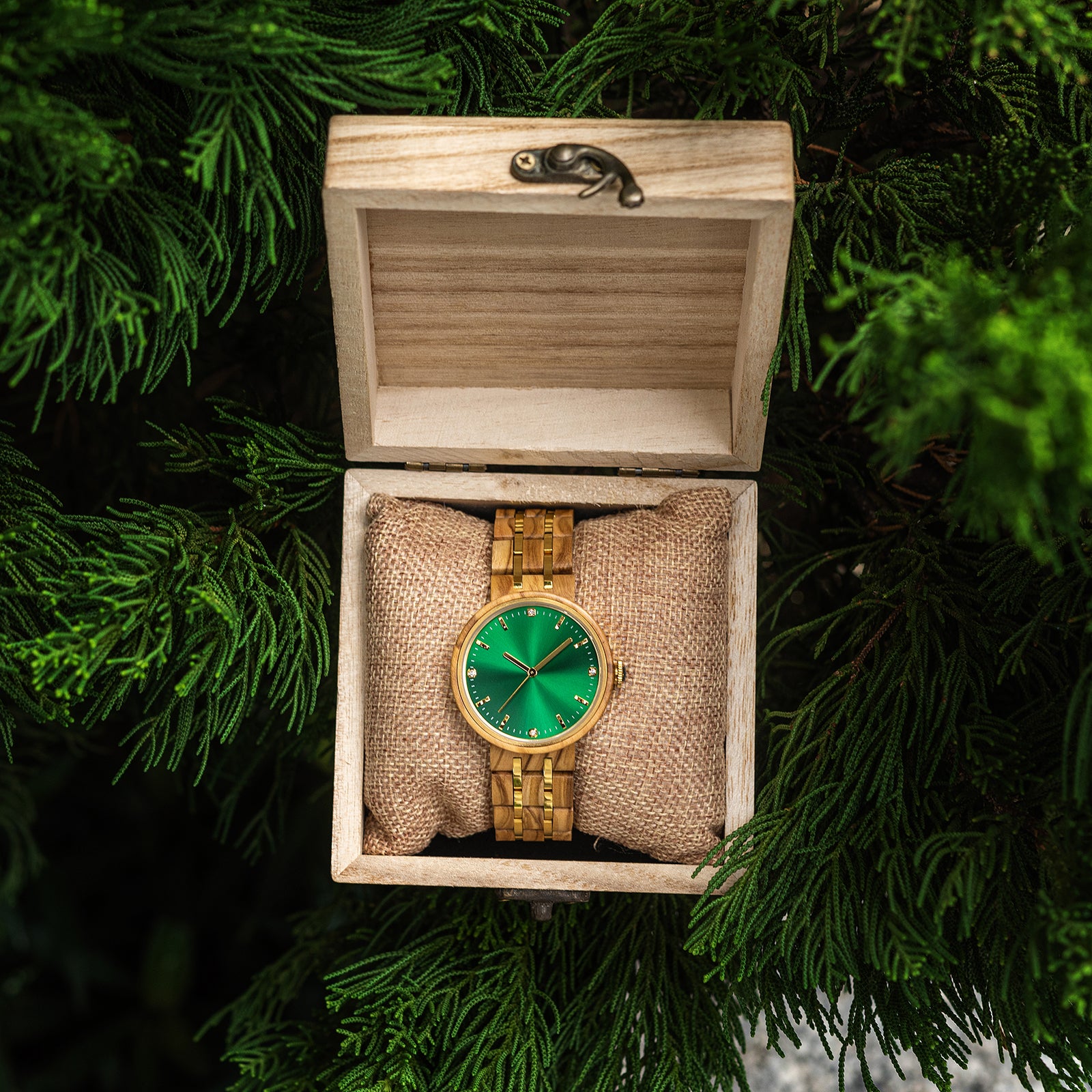 An elegant women's wooden watch featuring a green dial, gold-tone accents, diamond-like details, and a two-tone wood and metal band, presented in a wooden box with a burlap pillow amidst green foliage to highlight its luxurious and natural design.