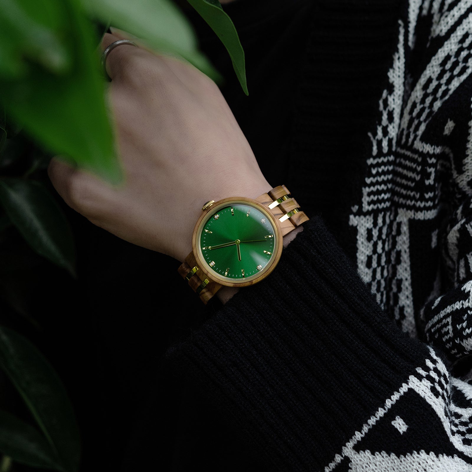 An elegant women's wooden watch featuring a green dial, gold-tone accents, diamond-like details, and a two-tone wood and metal band, showcased on a wrist against a backdrop of green foliage and black knitwear to highlight its luxurious and natural design.