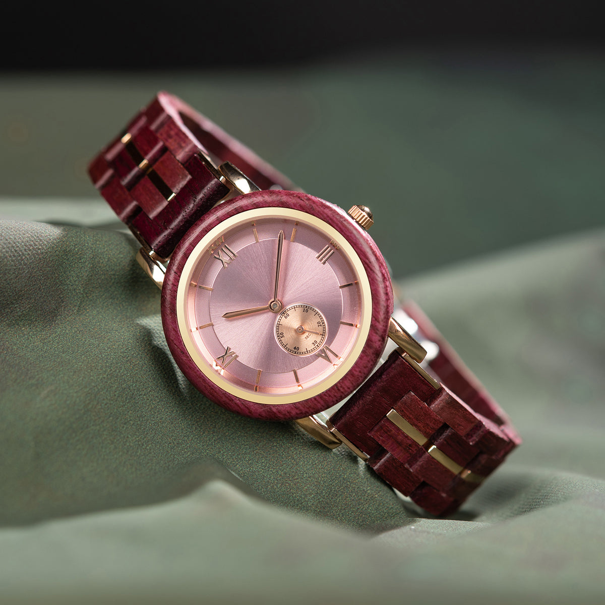 A stylish women's wooden watch featuring a pink dial, Roman numerals, sub-dial, and gold-accented details, presented against a green fabric background to highlight its sophisticated and elegant aesthetic.