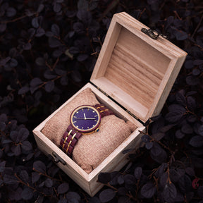 An elegant women's wooden watch featuring a purple dial, gold-tone accents, diamond-like details, and a two-tone wood and metal band, presented in a wooden box with a burlap pillow amidst dark foliage to highlight its luxurious and natural design.