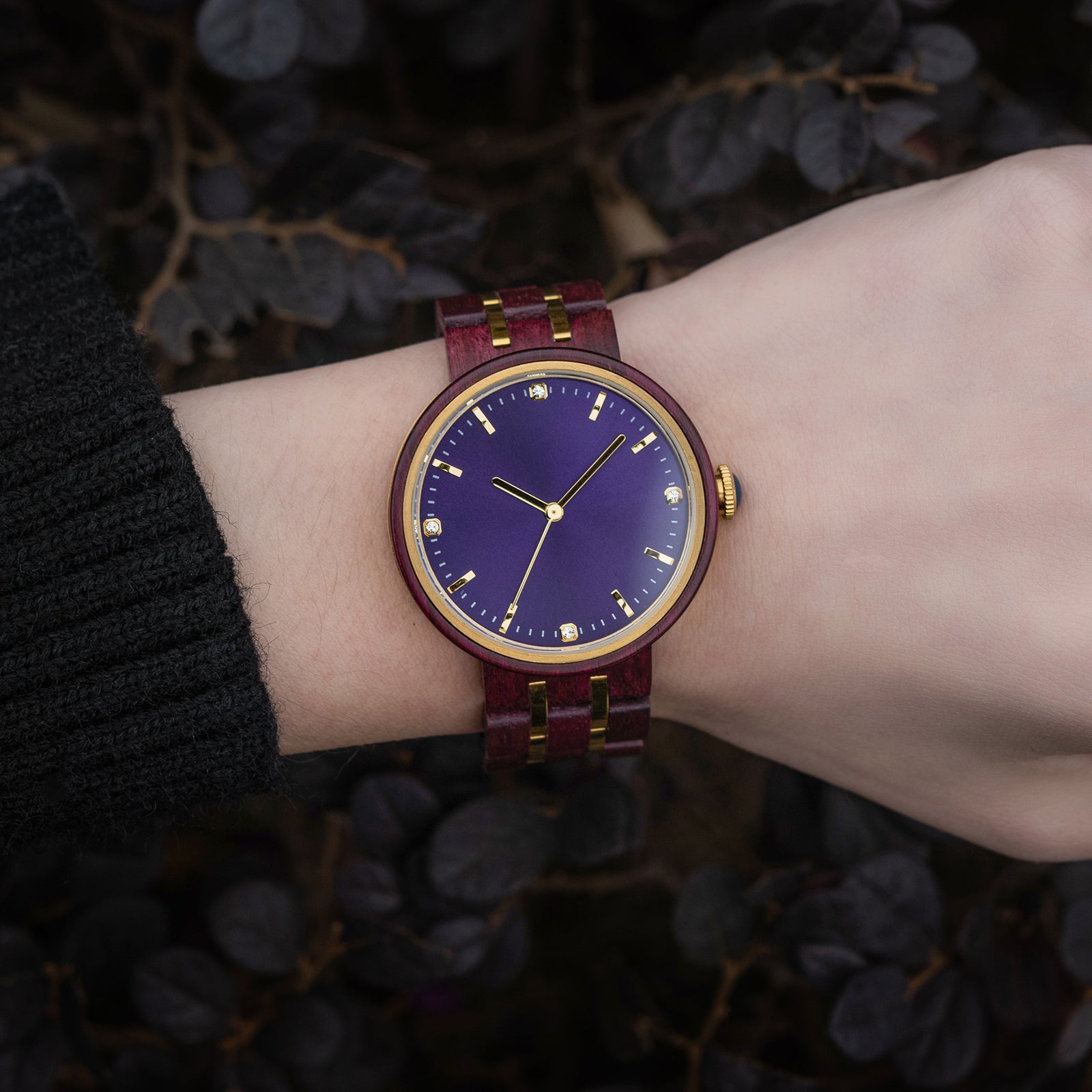 An elegant women's wooden watch featuring a purple dial, gold-tone accents, diamond-like details, and a two-tone wood and metal band, showcased on a wrist against a backdrop of dark foliage to highlight its luxurious and natural design.